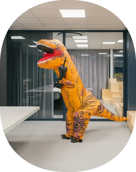 A person in a T-Rex costume with an inflated cell phone is standing in an office. They are pretending to be on a call.